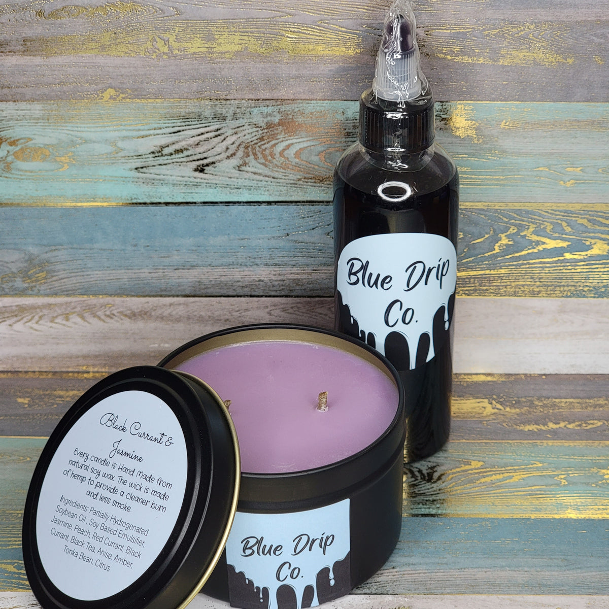A lavender scented candle in a black container with a lid and a scented oil bottle from Blue Drip Co on a wooden surface.