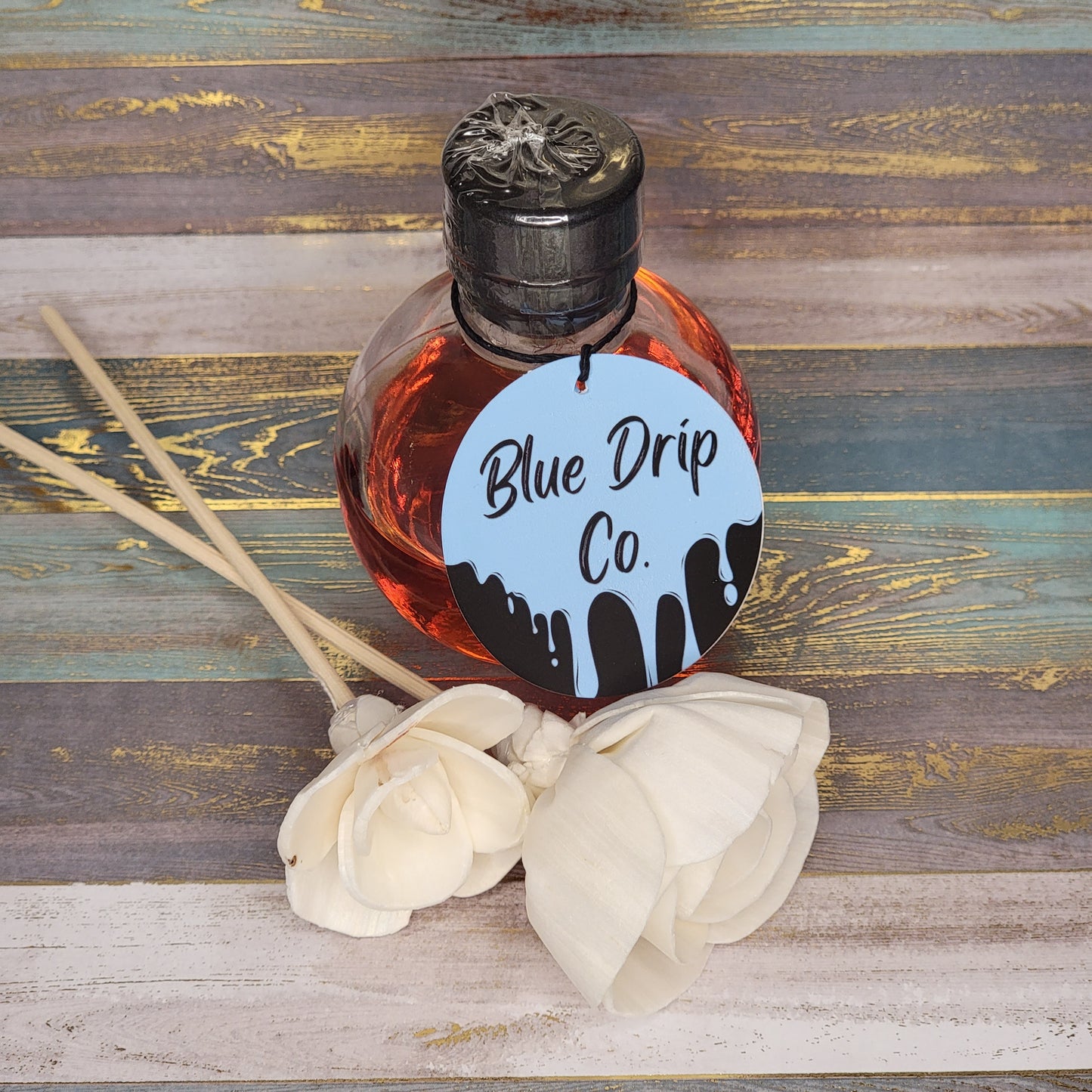 A decorative bottle labeled 'Blue Drip Co.' surrounded by white flowers on a textured wooden background.
