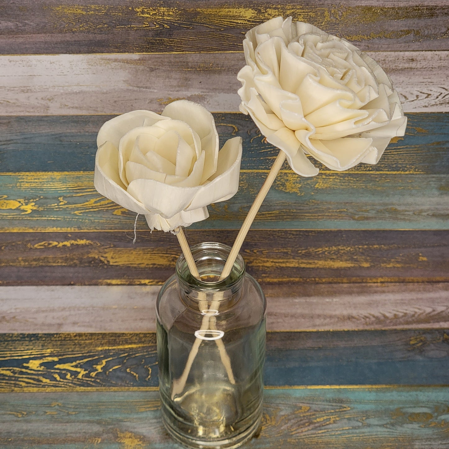 Two artificial cream-colored roses in a glass jar against a textured wooden background.