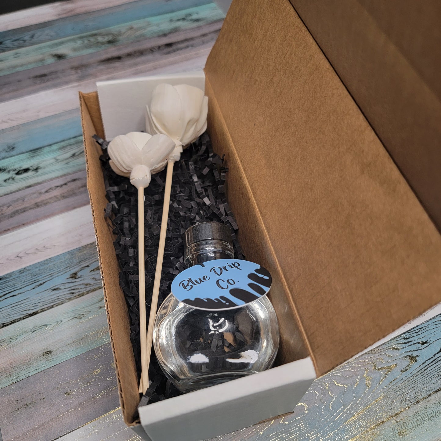 A packaged gift box containing a clear bottle labeled 'Blue Drip Co' and decorative flower sticks.