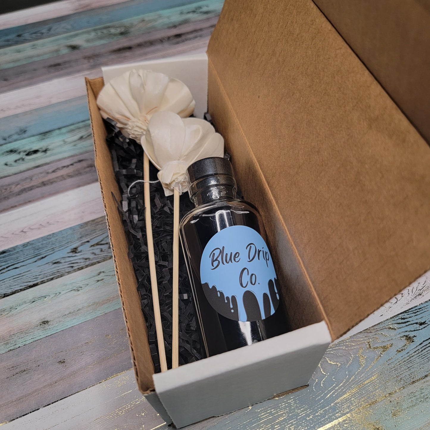 A bottle labeled 'Blue Drip Co.' with two white flower decorations, placed in a cardboard box.