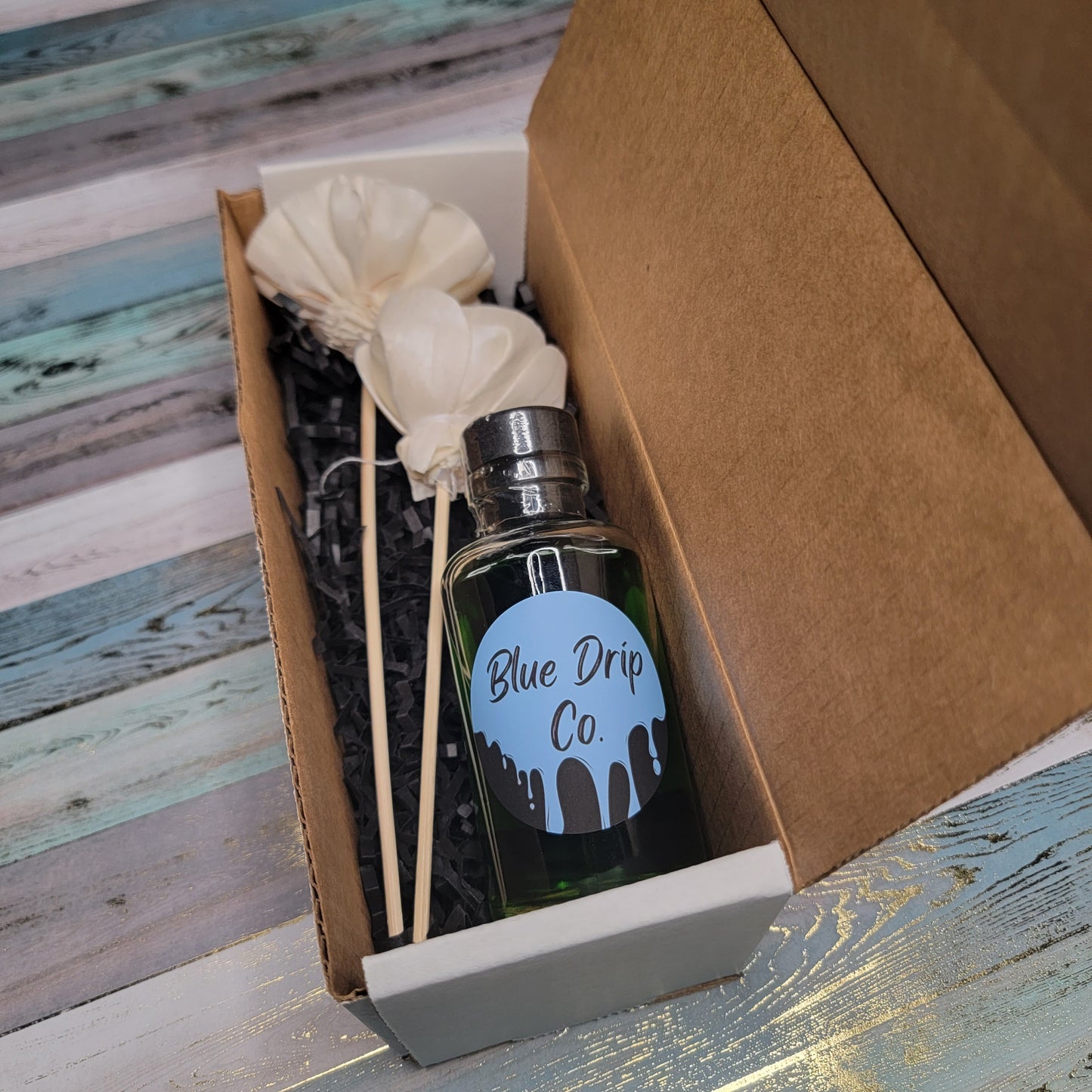 A bottle labeled 'Blue Drip Co.' placed in a box with decorative paper and white flower stems.