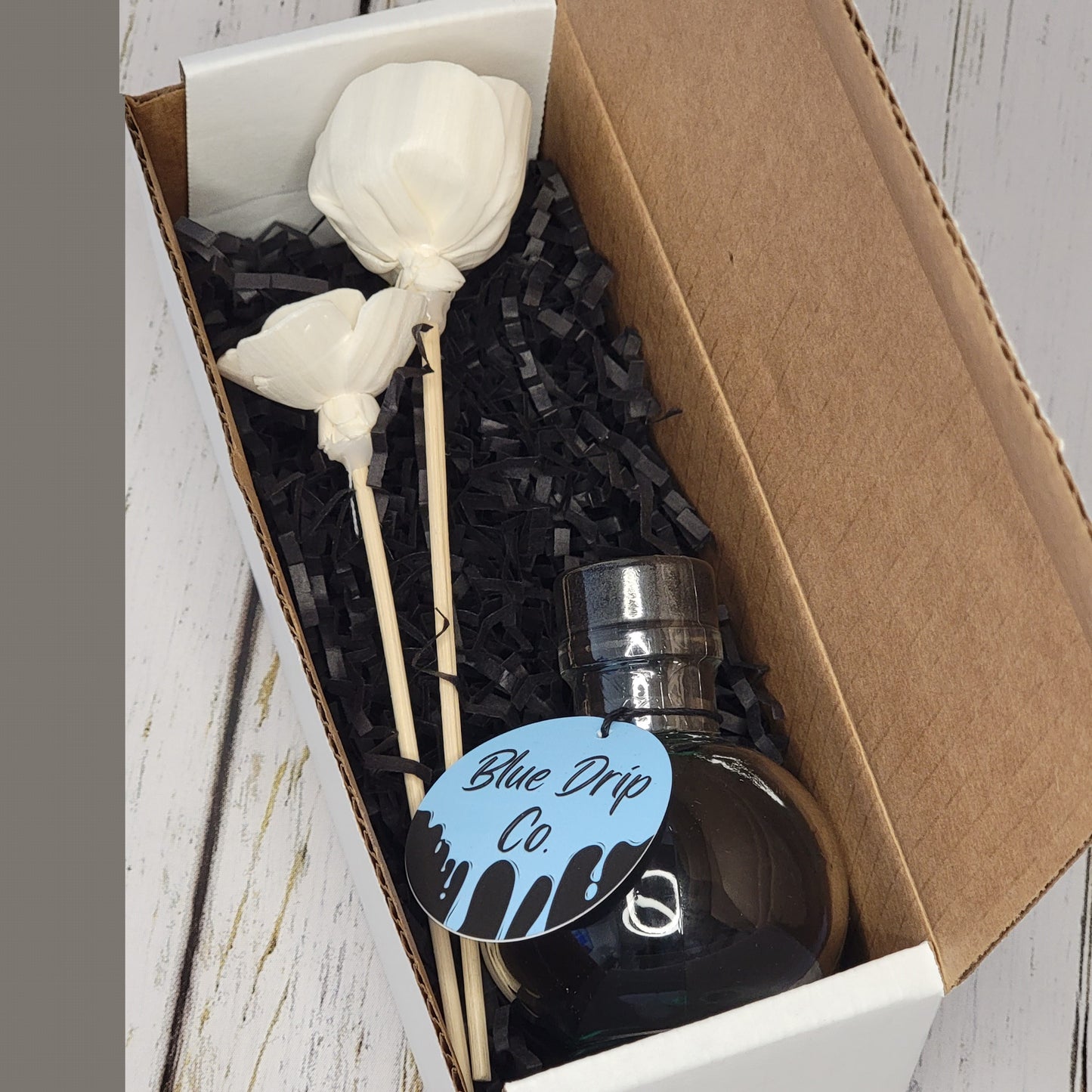 Gift box featuring a dark bottle labeled 'Blue Drip Co.' and two white decorative flowers on sticks, arranged with black shredded paper.