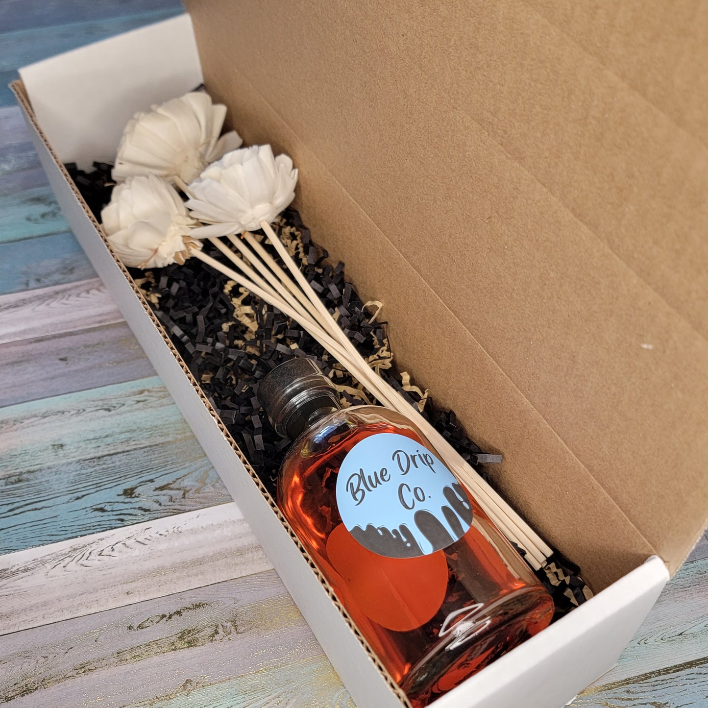 A decorative gift box containing a bottle labeled 'Blue Drip Co' alongside artificial flowers.