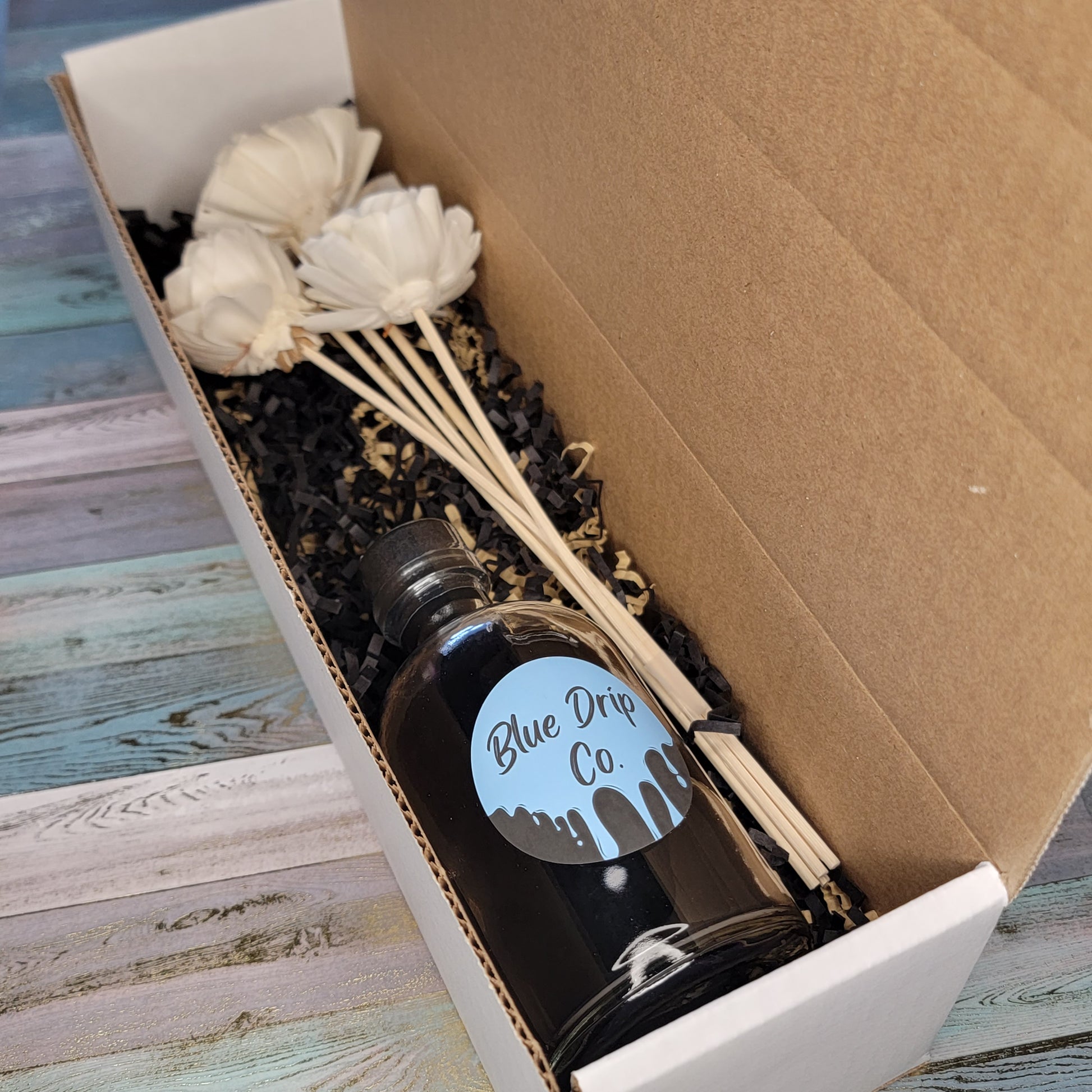 A gift box containing a bottle labeled 'Blue Drip Co' and decorative white flowers on sticks.