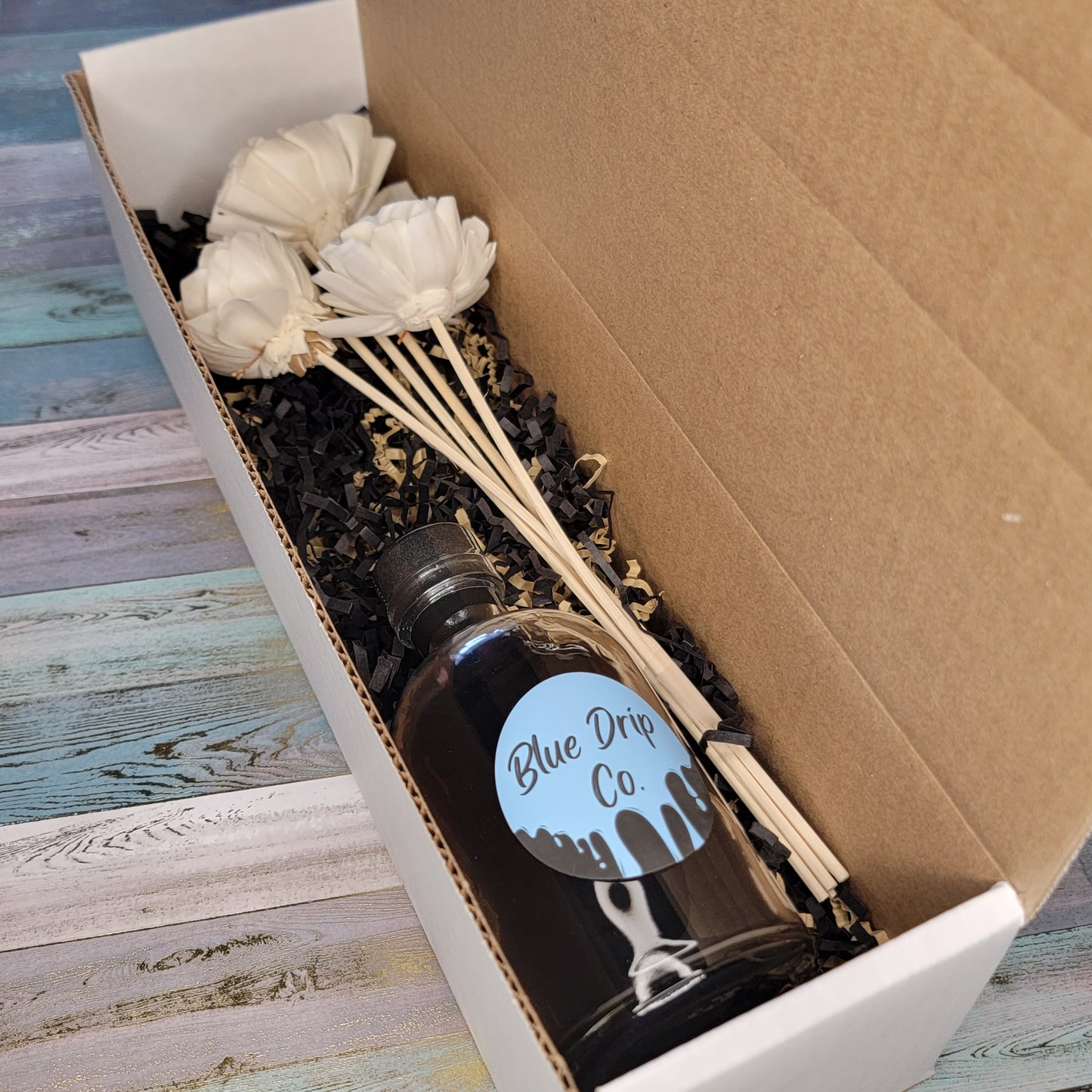 A gift box containing a bottle labeled 'Blue Drip Co.' and decorative white flowers on wooden sticks.