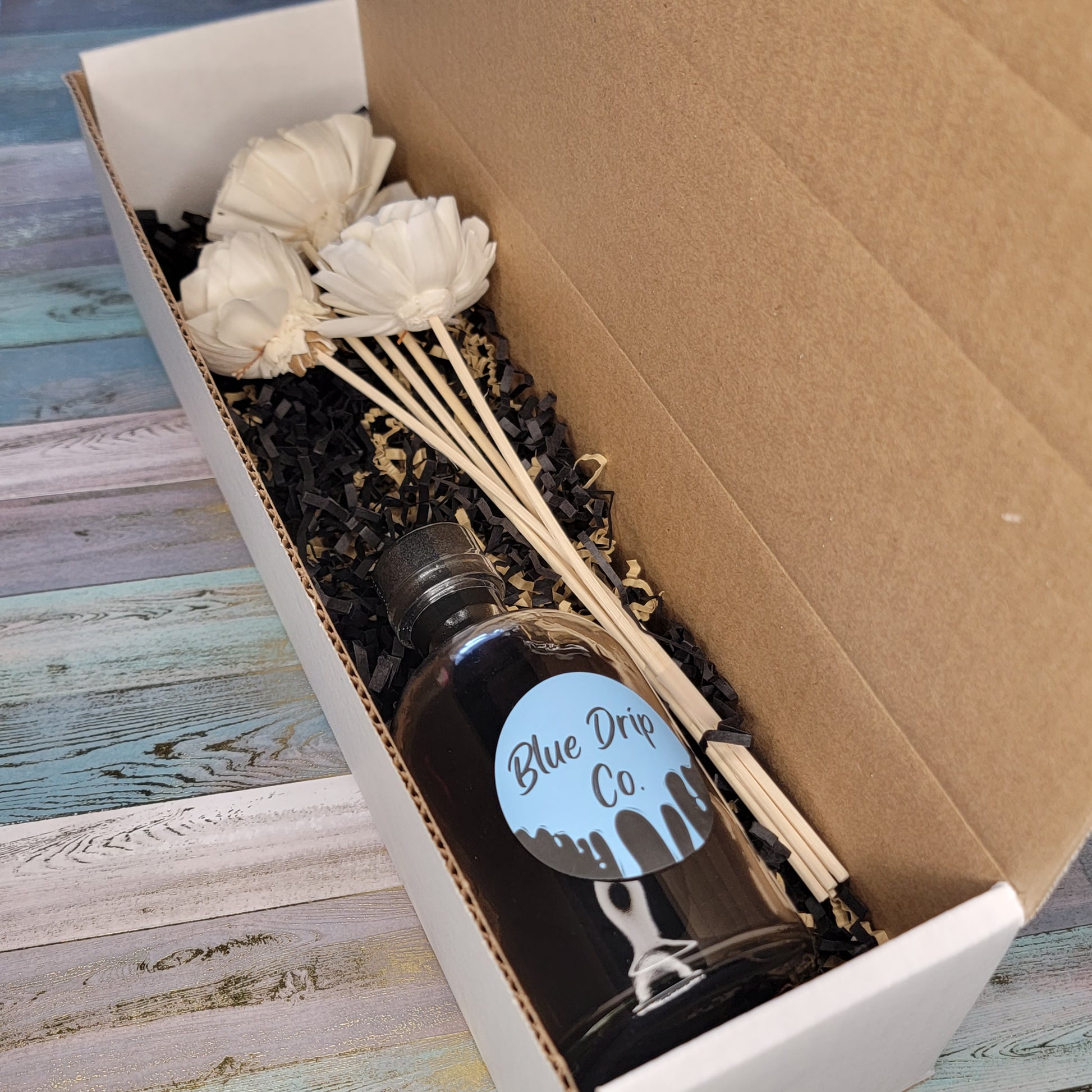A gift box containing a bottle labeled 'Blue Drip Co.' and decorative white flowers on wooden sticks.