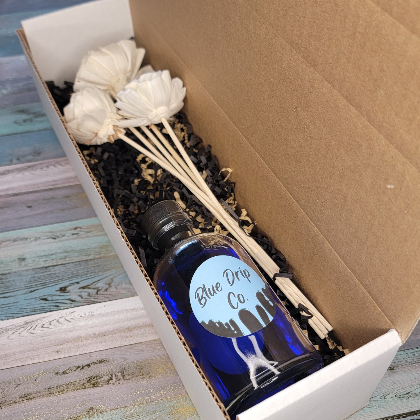 A blue bottle labeled 'Blue Drip Co.' surrounded by decorative fillers and white flower sticks in a brown box.