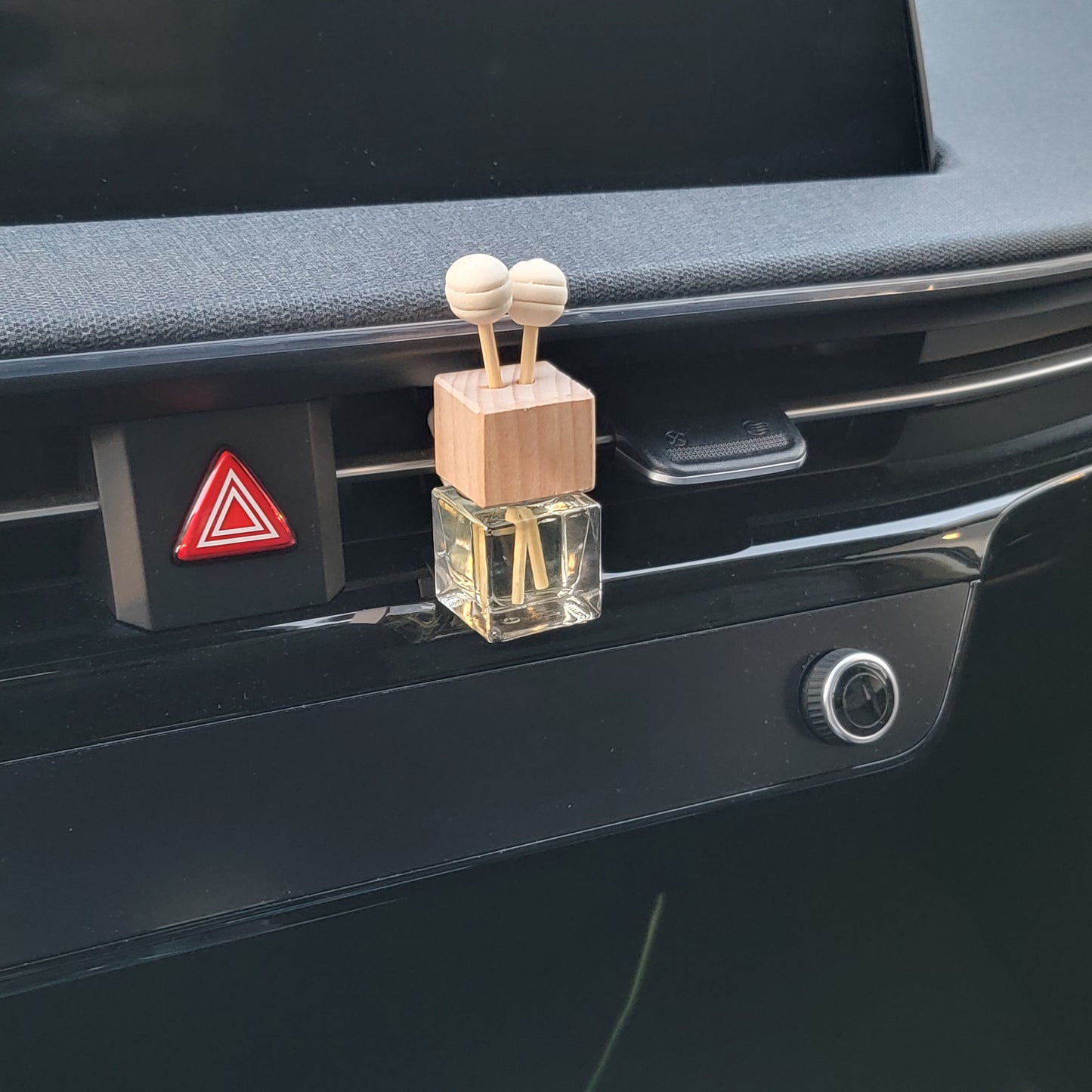 A small glass car air freshener with a wooden top and two diffuser sticks, placed near a hazard light button.
