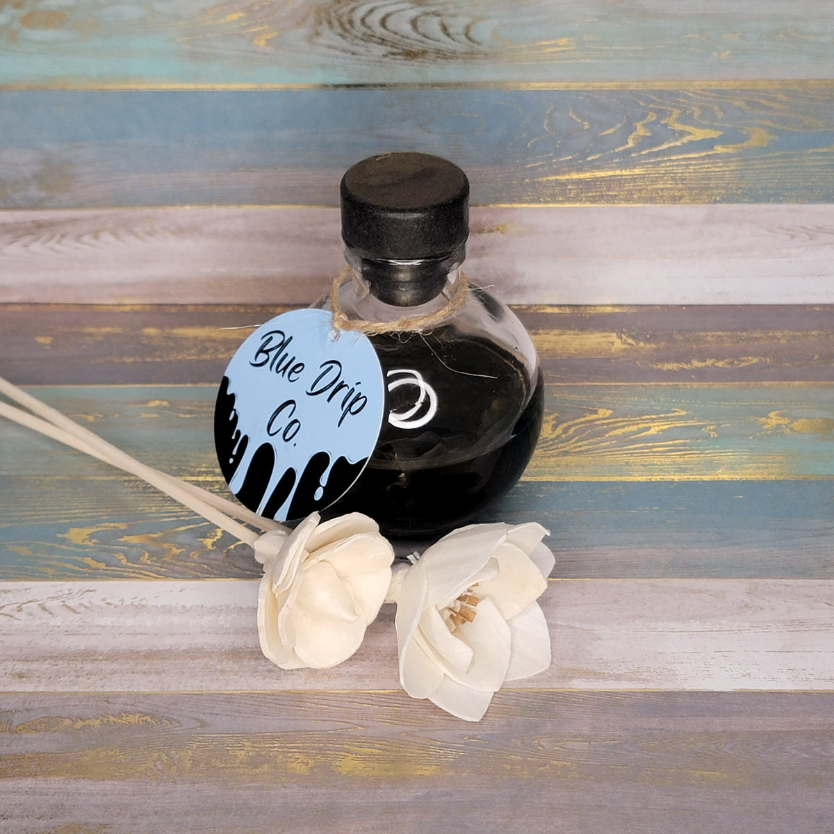 A glass bottle labeled 'Blue Drip Co.' containing dark liquid, accompanied by two white artificial flowers on a wooden surface.