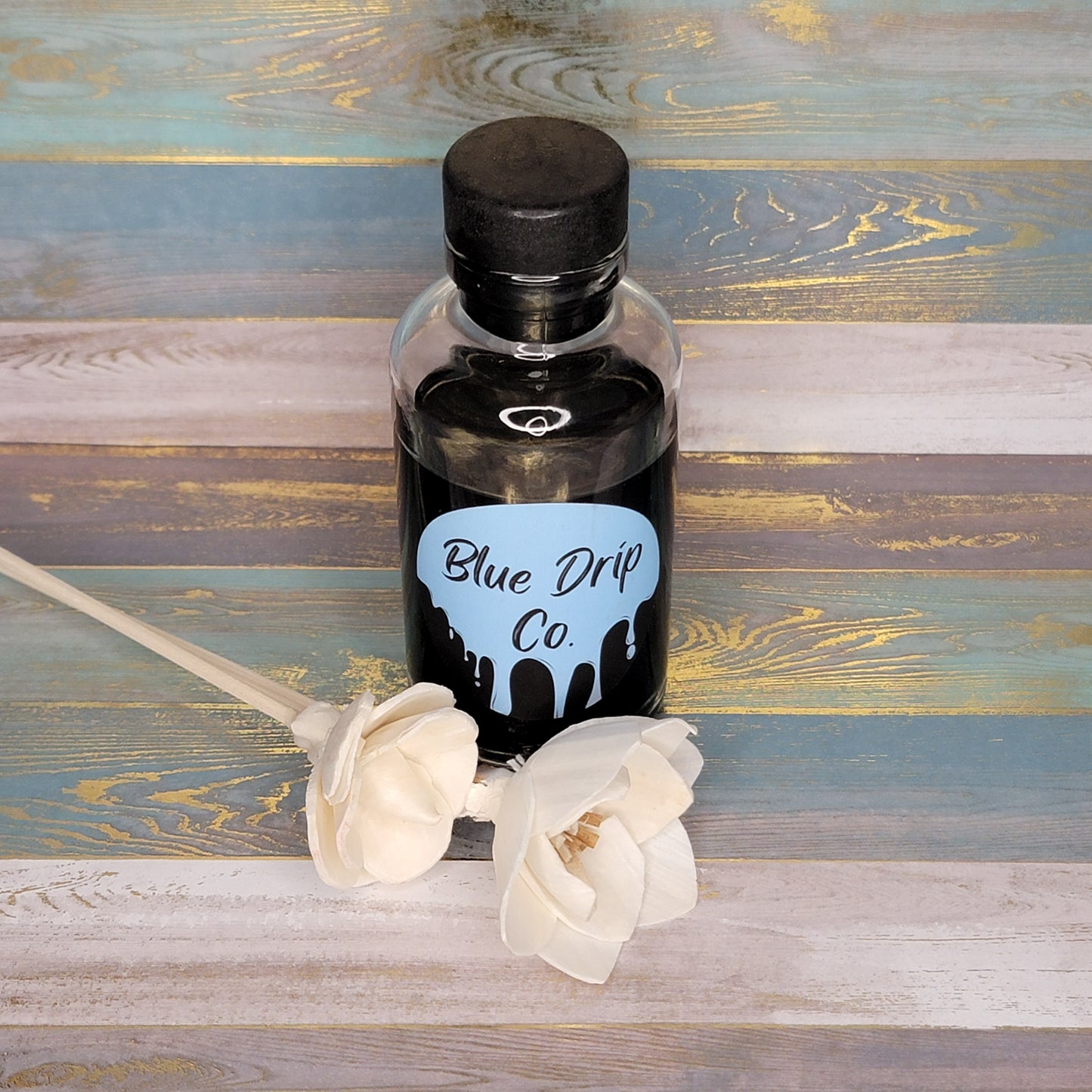A bottle labeled 'Blue Drip Co.' with a black liquid, accompanied by two white flowers on a wooden surface.