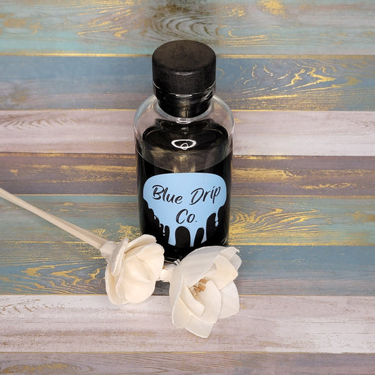 A bottle labeled 'Blue Drip Co.' with a black liquid, accompanied by two white flowers on a wooden surface.