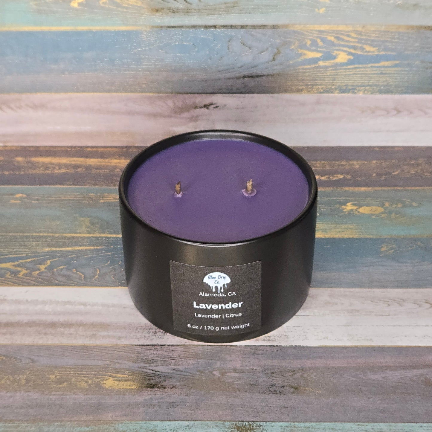 A round black candle container with lavender wax and two wicks on a colorful wooden background.