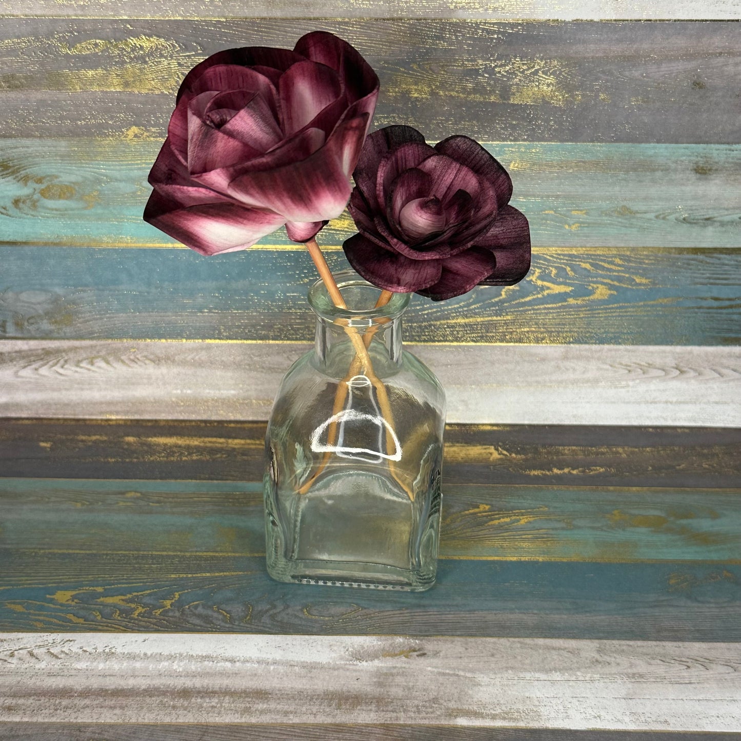 A glass vase holding two artificial purple roses against a rustic wooden background.