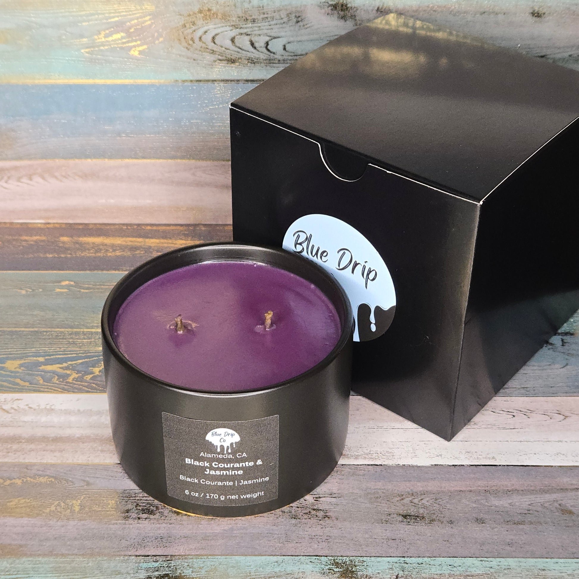 A round black candle with a purple wax and two wicks, next to its black box labeled 'Blue Drip'.