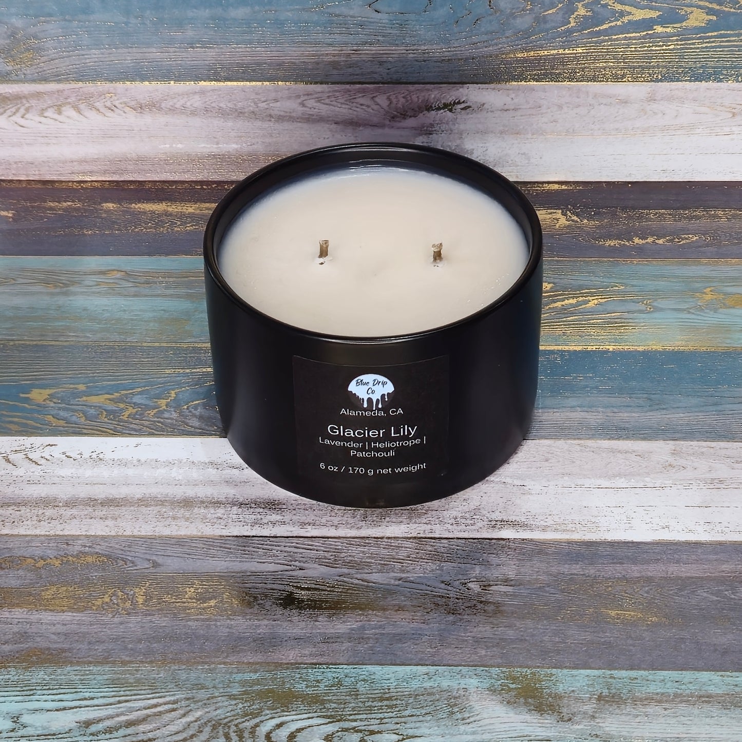 Black candle in a round container labeled 'Glacier Lily' on a wooden surface.