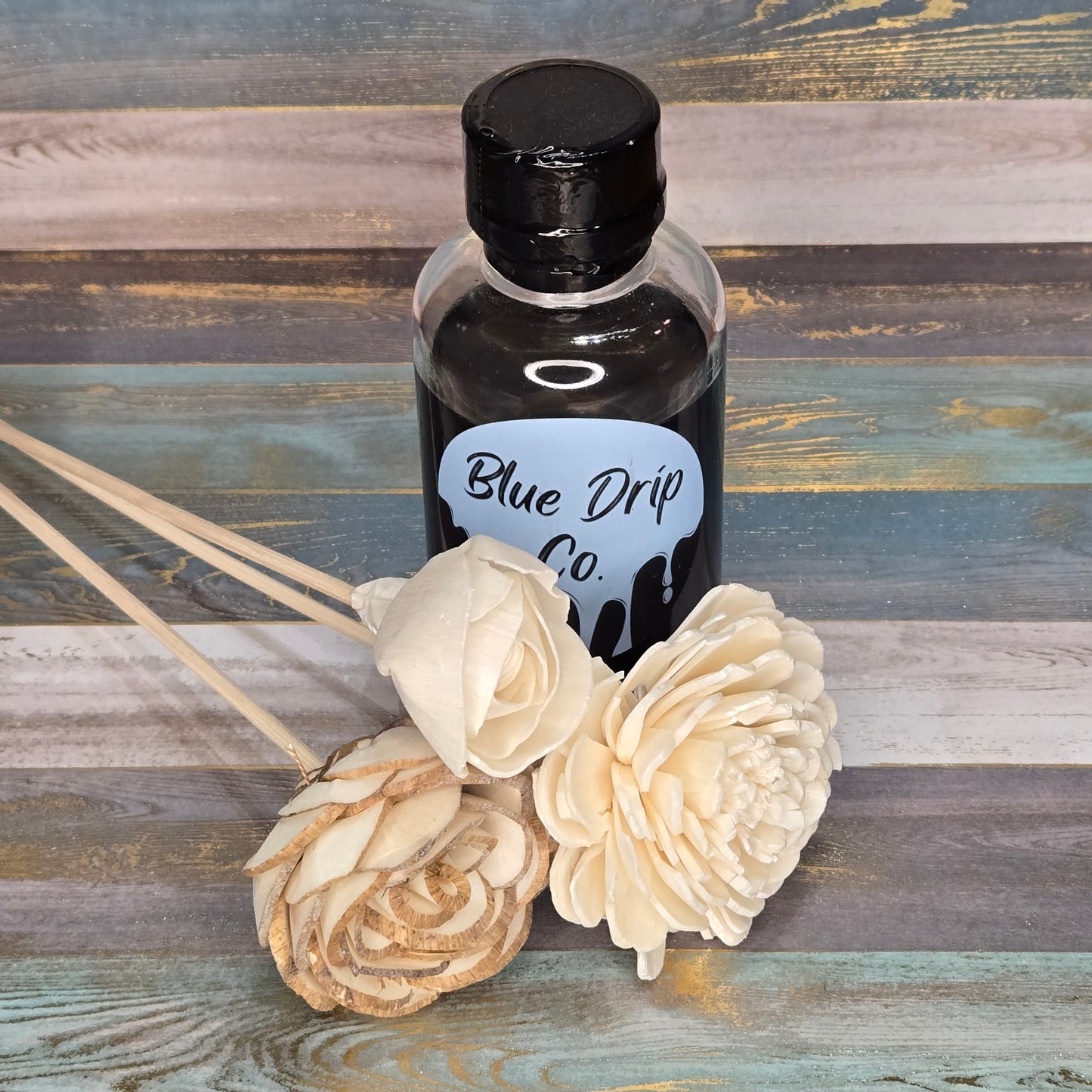 A glass bottle labeled 'Blue Drip Co.' with a black cap, surrounded by cream-colored decorative flowers and wooden sticks, set against a wooden background.